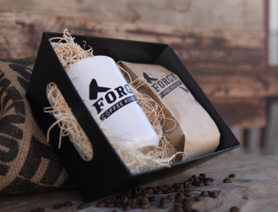 Image of a white ceramic mug and a bag of coffee resting in a black market basket filled with fiber fill.  It is sitting on a wood surface with a burlap coffee bag propping up the market basket.