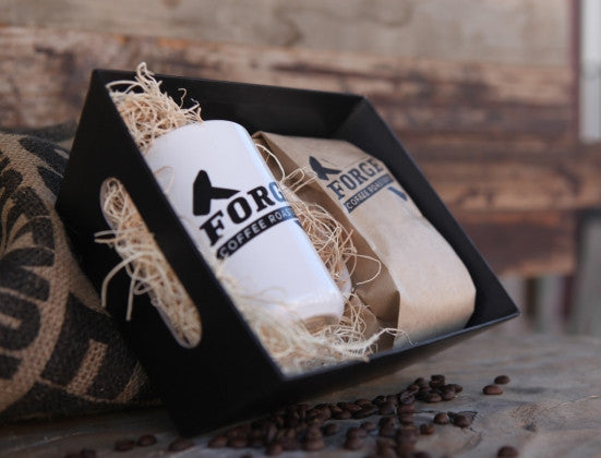 Image of a white ceramic mug and a bag of coffee resting in a black market basket filled with fiber fill. It is sitting on a wood surface with a burlap coffee bag propping up the market basket.